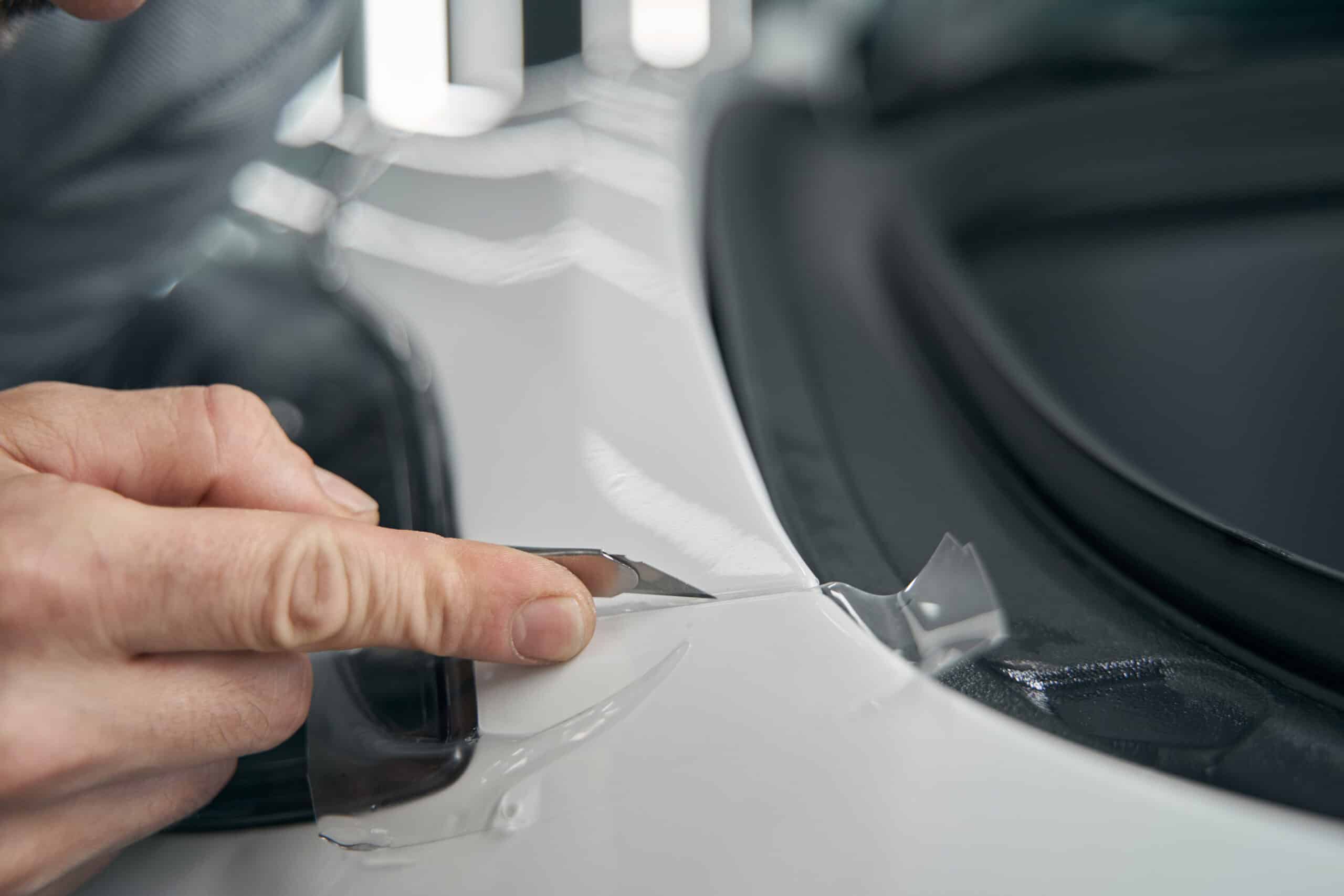 Nahaufnahme der Hand eines Fahrzeugaufbereiters, der mit einem Universalmesser überschüssige Klarsichtschutzfolie abschneidet.